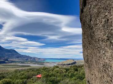 Dramatic lenticular clouds sweep across a vivid blue sky above a turquoise lake, lush green landscape, rugged mountains, and a lone red-roofed structure below.
