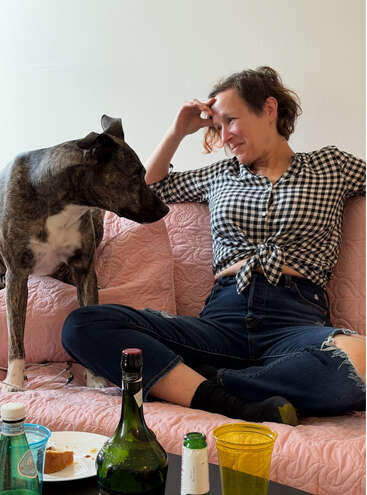 Uma mulher de camisa xadrez senta-se em um sofá rosa, compartilhando um momento descontraído e afetuoso com seu cão. Bebidas e comida estão sobre a mesa.