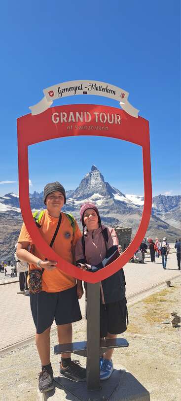 Two people stand beneath a “Grand Tour of Switzerland” frame at Gornergrat with the Matterhorn mountain in the background, surrounded by clear skies and other tourists.