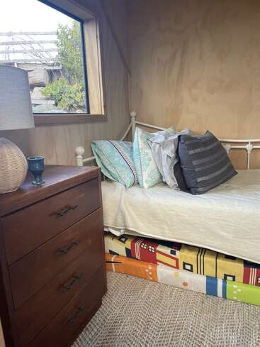 Un coin de chambre confortable avec un lit simple, des oreillers colorés, une commode en bois, une lampe et une fenêtre. Matelas ou coussins aux motifs audacieux rangés sous le lit.