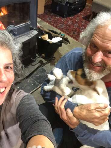 Un couple heureux est assis près d'un poêle à bois confortable, souriant chaleureusement, tandis que l'homme tient affectueusement un chien enjoué dans ses bras. Atmosphère confortable et accueillante.