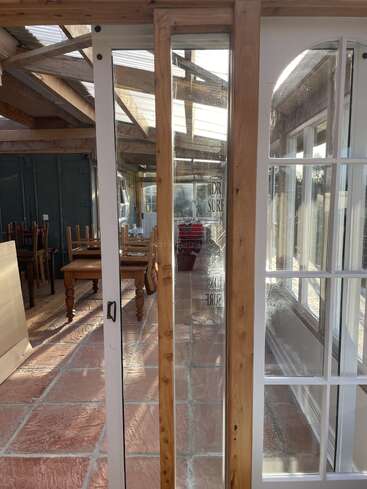 Un espace intérieur ensoleillé avec des sols en carreaux de terre cuite, des poutres en bois, des tables et des chaises, de multiples portes et fenêtres en verre reflétant la lumière et une structure en bois ouverte.
