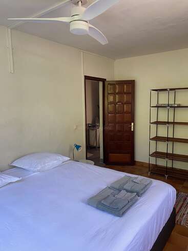 Esta imagen muestra un dormitorio sencillo con una cama grande, ropa de cama blanca, toallas dobladas, ventilador de techo, puerta abierta, estantería, lámpara y suelo de baldosas.