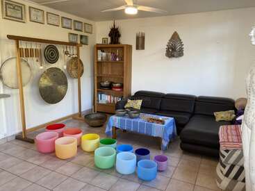 Esta acogedora sala cuenta con coloridos cuencos tibetanos de cristal, gongs, cuencos tibetanos, un sofá de cuero, una estantería, certificados en la pared y un ambiente sereno y meditativo.