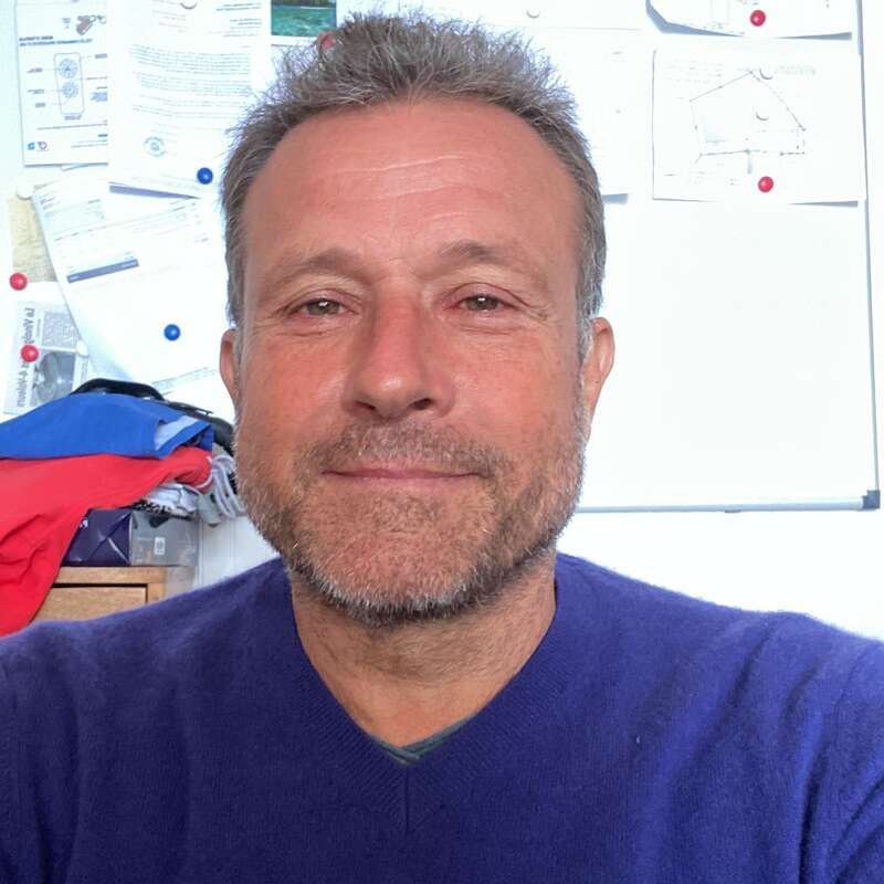 Un hombre de pelo corto y barba recortada sonríe amablemente. Lleva un jersey azul y está sentado delante de una pizarra desordenada.