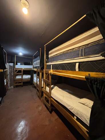 Esta imagem mostra um quarto mal iluminado com vários beliches de madeira alinhados, colchões e roupas de cama simples e cortinas para privacidade, típico de um dormitório de albergue.