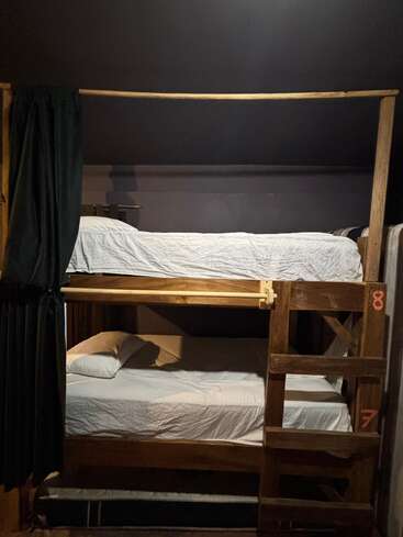 Esta imagem mostra um beliche de madeira com lençóis e travesseiros brancos. Uma escada dá acesso ao beliche superior, e cortinas escuras oferecem alguma privacidade.