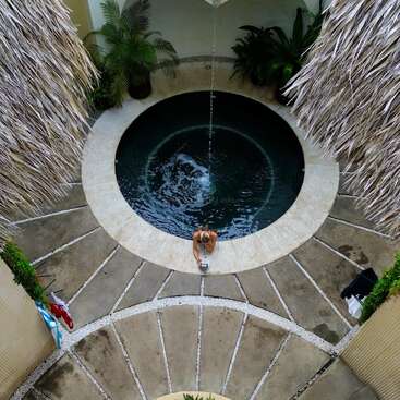 Vista aérea de uma piscina redonda com uma pessoa sentada na borda, cercada por plantas tropicais e telhados de palha, com água entrando na piscina.