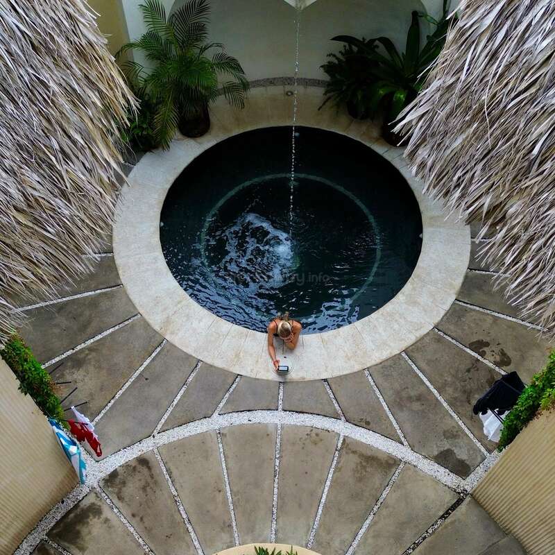 Vista aérea de uma piscina redonda com uma pessoa sentada na borda, cercada por plantas tropicais e telhados de palha, com água entrando na piscina.