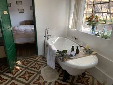 Une salle de bain confortable avec une baignoire sur pieds, un plateau en bois contenant des serviettes, du vin et des fleurs, un sol carrelé à motifs, une porte verte et la lumière du soleil qui pénètre par de grandes fenêtres.