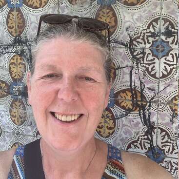 Una mujer sonríe cálidamente mientras se hace un selfie frente a una pared de azulejos coloridos con bonitos diseños. Lleva gafas de sol en la cabeza y un top sin mangas a rayas.