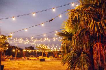 Una escena al aire libre al atardecer, con guirnaldas de luces colgando del cielo, que iluminan una zona de arena con palmeras y asientos dispersos, creando un ambiente cálido, festivo y acogedor.