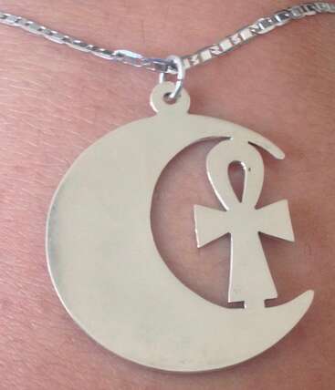 A silver necklace features a pendant combining a crescent moon and an ankh symbol, resting against skin, symbolizing a blend of ancient Egyptian and lunar motifs.