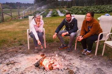 Three people sit in chairs around a campfire, roasting marshmallows. Behind them, there’s a lush green garden and sprinklers watering the rows of crops.