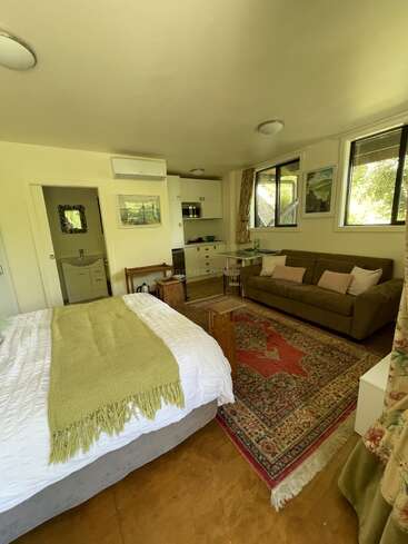 Dieses gemütliche Studiozimmer verfügt über ein Bett, eine grüne Decke, ein braunes Sofa, einen Vintage-Teppich, eine kleine Küche, einen Schreibtisch, Wandkunst, große Fenster und viel Licht.