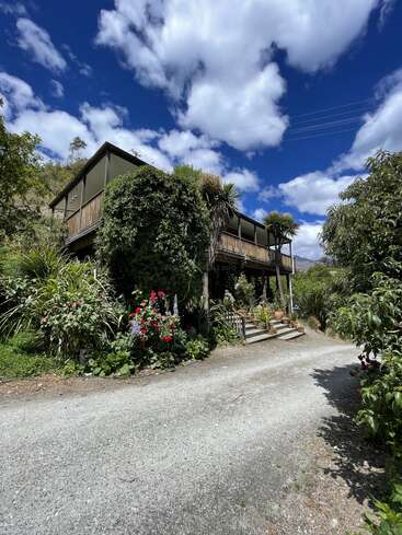 Ein rustikales Holzhaus, umgeben von üppigem Grün und bunten Blumen, unter einem strahlend blauen Himmel mit flauschigen weißen Wolken, entlang eines friedlichen Kieswegs.