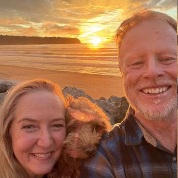 Ein lächelndes Paar und ihr flauschiger Hund machen ein Selfie am Strand bei Sonnenuntergang. Das goldene Sonnenlicht, die ruhigen Wellen und die felsige Küste schaffen eine warme, fröhliche Szene.