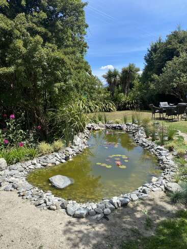 Ein kleiner Gartenteich ist von Steinen umgeben, auf denen Seerosen schwimmen. Üppig grüne Bäume, Sträucher, rosa Blumen und Sitzgelegenheiten im Freien schaffen eine friedliche Atmosphäre.
