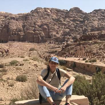 The image depicts a woman sitting on a rock in front of a desert landscape with ancient ruins and a large rocky mountain in the background.