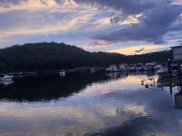 Uma marina tranquila ao pôr do sol com barcos ancorados na costa, águas calmas refletindo o céu colorido, cercada por colinas arborizadas e beleza natural serena.