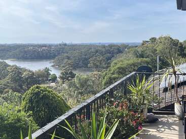 Uma varanda verde e exuberante, repleta de plantas, tem vista para um lago sereno, cercado por uma densa floresta sob um céu claro, proporcionando uma vista natural panorâmica e tranquila.