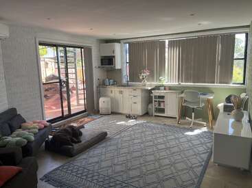 Uma sala de estar bem iluminada com janelas grandes, uma pequena cozinha, sofá, escrivaninha e um cachorro relaxando em uma cama. Ambiente aconchegante, organizado e convidativo.