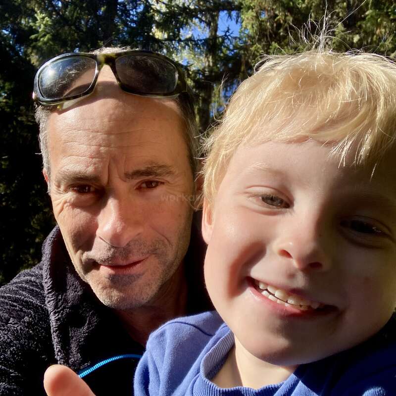 Un homme et un jeune garçon sourient chaleureusement pour un selfie en gros plan à l'extérieur, entourés d'arbres verts et de soleil, capturant un moment joyeux et affectueux ensemble.