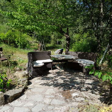 Auf einer Steinterrasse befindet sich eine gemütliche Sitzecke im Freien mit gepolsterten Bänken und einem runden Tisch, umgeben von üppig grünen Bäumen und Gartenpflanzen.
