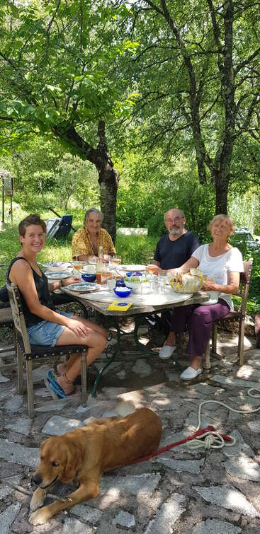 Vier Leute sitzen an einem Tisch im Freien und genießen gemeinsam eine Mahlzeit unter laubigen Bäumen, während sich ein goldfarbener Hund auf der Steinterrasse davor ausruht.