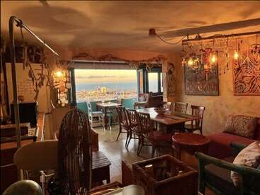 Un espace de vie confortable et éclectique avec un éclairage chaleureux, des meubles en bois et une décoration artistique donne sur un port de plaisance pittoresque et l'océan au coucher du soleil à travers de grandes fenêtres ouvertes.