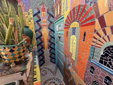 Cette image vibrante montre une pièce étroite avec des murs en mosaïque colorée, une porte bleu clair, des plantes serpentines en pot et des motifs géométriques complexes dans tout l'espace.