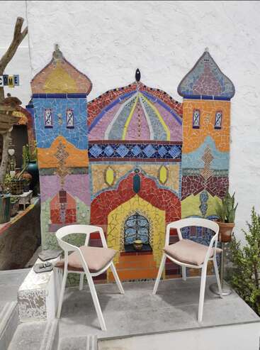 Deux chaises blanches avec des coussins beiges font face à un mur en mosaïque colorée, représentant un château orné. Des plantes en pot et une atmosphère confortable et accueillante complètent cette scène charmante.