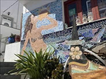 Une mosaïque murale colorée représente une sirène buvant dans un coquillage. Entourée de plantes, de lanternes et de décorations nautiques, cette scène vibrante illumine la façade du bâtiment.