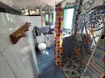 Cette salle de bains pleine de vie présente des carreaux de mosaïque, des toilettes blanches, un sol bleu, un rideau de douche à pois coloré, un portemanteau en bois, des miroirs et une fenêtre à cadre vert.
