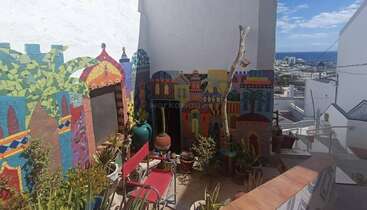 Un patio animé présente des mosaïques murales colorées représentant des bâtiments et des palmiers, avec diverses plantes en pot, des chaises rouges, des pots en céramique et une vue pittoresque sur la côte.