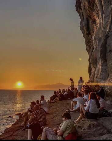 Un groupe de personnes se détend sur des falaises rocheuses, en regardant un magnifique coucher de soleil sur l'océan. Le ciel s'illumine d'orange, créant une atmosphère sereine et paisible.