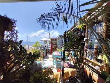 Un patio extérieur vibrant et éclectique rempli de plantes luxuriantes, de meubles colorés, d'œuvres d'art et de décorations de plage, surplombant un ciel bleu vif et l'horizon de l'océan.