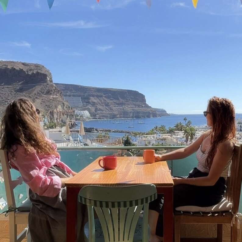 Deux femmes sont assises à une table avec des tasses de café, profitant d'une vue côtière ensoleillée de falaises, de mer bleue, de bateaux et d'une ville pittoresque de bord de mer sous des drapeaux de fête.