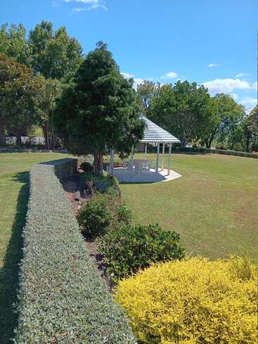 A peaceful garden scene with manicured hedges, colorful bushes, and a white gazebo under a clear blue sky, surrounded by lush green trees and grass.