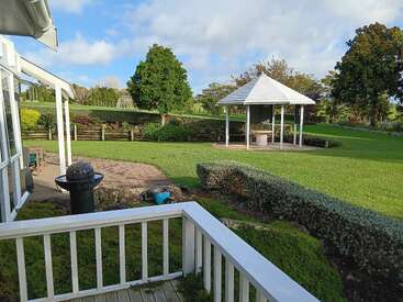 A peaceful garden scene with a white gazebo, green grass, trimmed hedges, trees, a patio area, and a white-fenced porch under a partly cloudy sky.