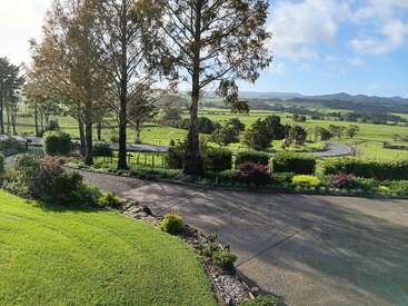 A serene countryside scene with lush green grass, trees, well-maintained gardens, a winding road, and distant hills under a partly cloudy blue sky. Peaceful and inviting.
