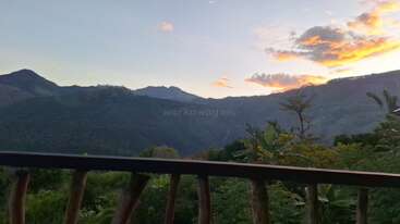 A beautiful mountain landscape at sunset, with lush green vegetation in the foreground, wooden railing, and dramatic clouds illuminated by the fading sunlight in the sky.