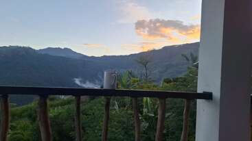 A white mug sits on a wooden railing, overlooking lush green hills and distant mountains at sunrise, with soft clouds glowing in the colorful sky.
