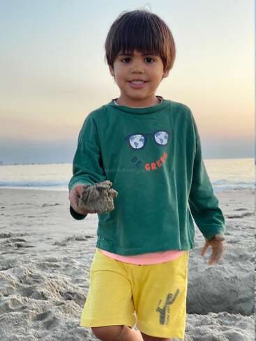 Ein lächelnder Junge steht bei Sonnenuntergang an einem Sandstrand und hält einen Sandklumpen in der Hand. Er trägt ein grünes Hemd und gelbe Shorts und genießt seine Spielzeit.