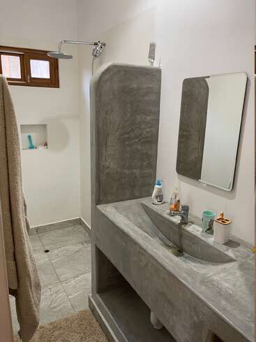 Dieses minimalistische Badezimmer verfügt über ein Waschbecken aus Beton, einen grau gefliesten Boden, eine wandmontierte Dusche, einen rechteckigen Spiegel, ein kleines Fenster, ein Handtuch, Toilettenartikel und eine eingebaute Duschablage.