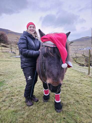 Una mujer sonriente, vestida con una chaqueta de invierno negra, está de pie junto a un poni marrón. El poni lleva un gorro rojo de Papá Noel y calentadores festivos en un prado.