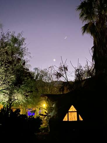 Uma cena noturna com árvores, uma lua brilhante e uma janela triangular iluminada em uma casa de campo escura. Luzes amarelas e azuis suaves realçam a atmosfera pacífica.
