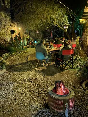 Um ambiente aconchegante ao ar livre à noite, onde as pessoas se sentam em torno de mesas próximas a uma fogueira, desfrutando de bebidas, comida e música ao vivo sob árvores com iluminação quente.