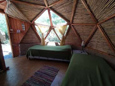 Este é um quarto aconchegante em forma de domo geodésico com duas camas de solteiro, cobertores verdes, paredes de madeira e bambu, janelas grandes, cortinas, uma lâmpada e um tapete colorido.
