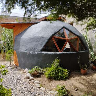 Esta imagem mostra uma pequena casa de adobe em forma de cúpula com janelas geométricas, cercada por plantas e cascalho, combinando materiais de construção naturais com um ambiente externo tranquilo.
